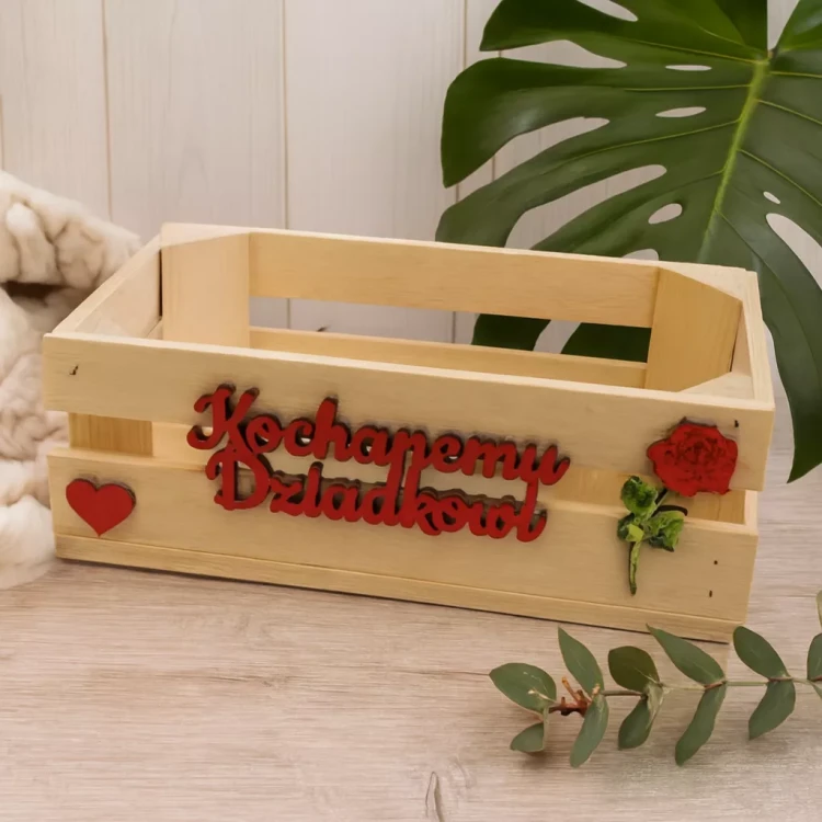Drewniana skrzynka z napisem „Kochanemu Dziadkowi”, ozdobiona czerwonym sercem, różą i zielonymi listkami. Ręcznie wykonana dekoracja idealna na prezent z okazji Dnia Dziadka lub urodzin, w ciepłej, domowej aranżacji.