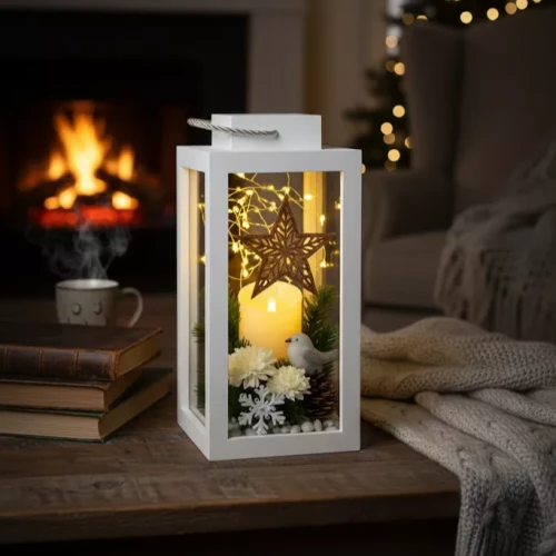 Biały lampion z dekoracją świąteczną: świeca, gwiazda z wikliny, sztuczny śnieg, kwiaty, szyszki, zielone gałązki i figurka ptaszka. W tle kominek, koc, choinka i kubek, przytulna, zimowa atmosfera.