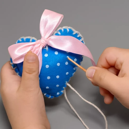 Ręce dziecka zszywają niebieskie filcowe serduszko w białe kropki plastikową igłą z białą włóczką. Serduszko ozdobione jest różową kokardą, a całość znajduje się na neutralnym tle, ukazując proces rękodzielniczy z bliska.