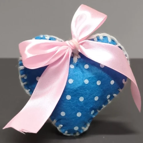 Ręcznie wykonana ozdoba w kształcie serca z niebieskiego filcu w białe kropki, obszyta białą nicią. U góry różowa satynowa kokardka. Tło neutralne, szare, podkreśla kolorystykę i detale dekoracji.