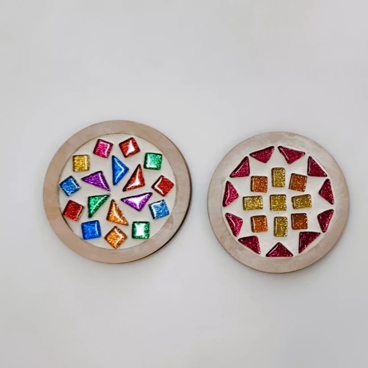 Dwie okrągłe drewniane podstawki pod kubek z kolorowymi, błyszczącymi figurami geometrycznymi. Lewy z nieregularnym układem kształtów w różnych kolorach, prawy z symetrycznym wzorem z czerwonych trójkątów i żółto-pomarańczowych kwadratów.