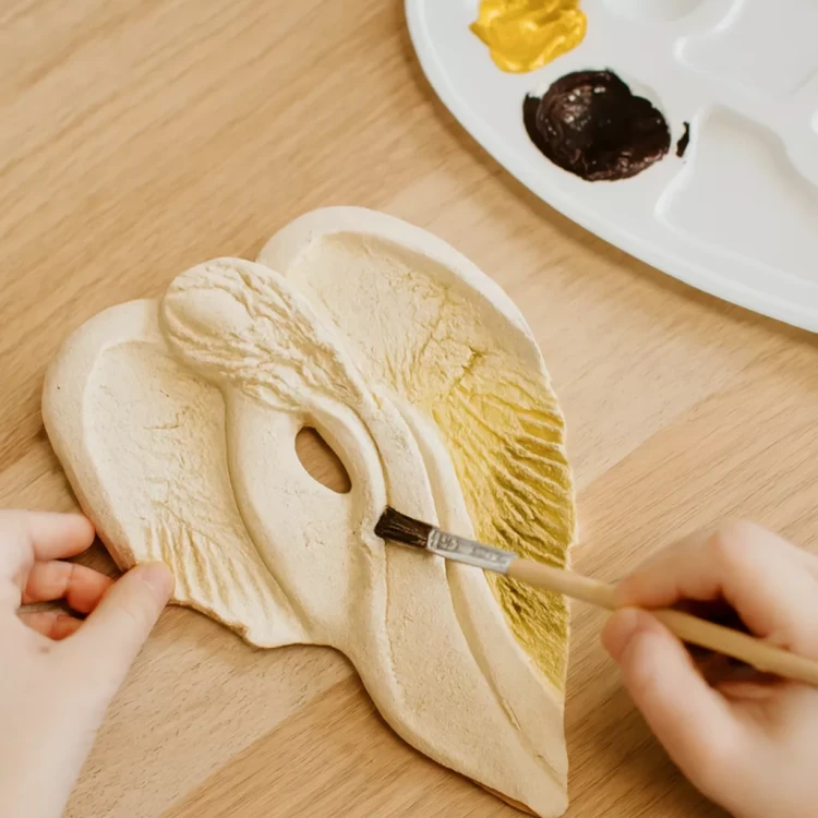 Osoba maluje gipsowego anioła o spokojnej twarzy i rozłożystych skrzydłach. Pędzlem nanosi żółtą farbę na skrzydło. Na stole widać paletę z farbami i drewniane podłoże, co podkreśla rękodzielniczy charakter sceny.