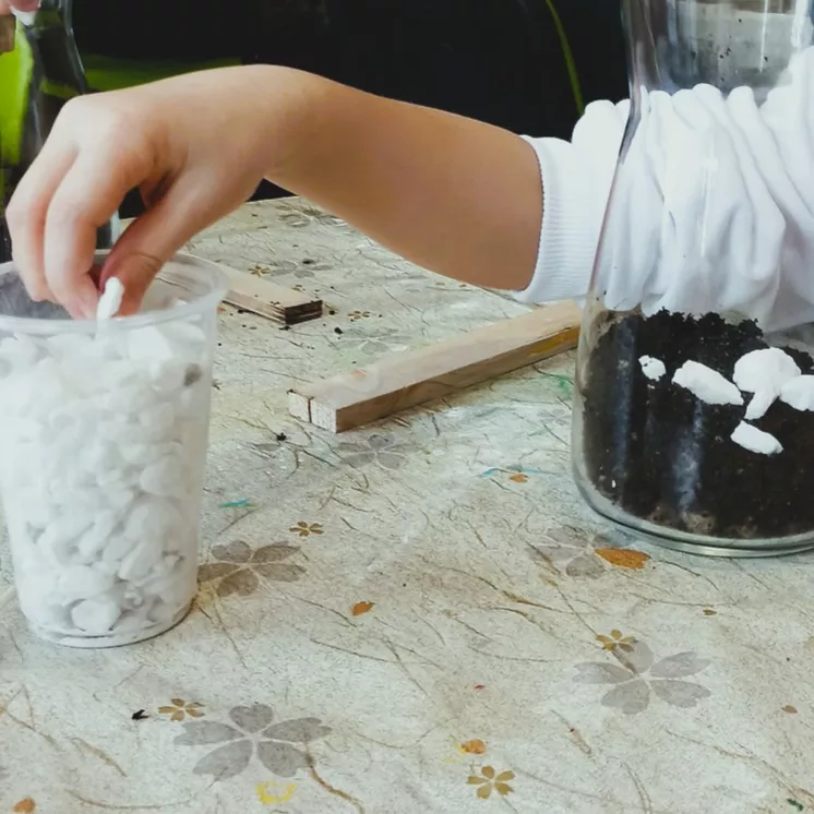 Dziecko wsypuje  białe kamyki z kubka do szklanego naczynia z ziemią. Wokół rozrzucone patyczki i materiały, sugerujące warsztaty edukacyjne i tworzenie miniaturowego ogrodu.