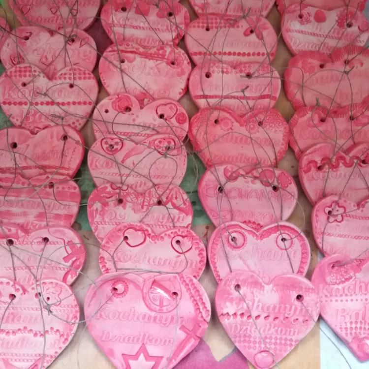 Ręcznie wykonane różowe ceramiczne serduszka z tłoczonymi wzorami i napisem, ułożone w rzędach. Każde ma otwory z przewleczonym sznurkiem do zawieszenia dekoracje na każdą okazję, pełne ciepła i różnorodnych detali.