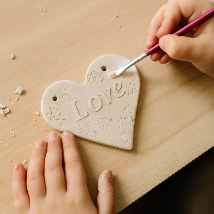 Dziecięce dłonie ozdabiają serduszko z gliny z napisem „Love”. Jedna ręka maluje pędzelkiem, druga opiera się o stół. Serce ma otwory do zawieszenia i delikatne tłoczenia. Na stole widoczne są drobinki gliny. Jest to scena twórczej pracy ręcznej.