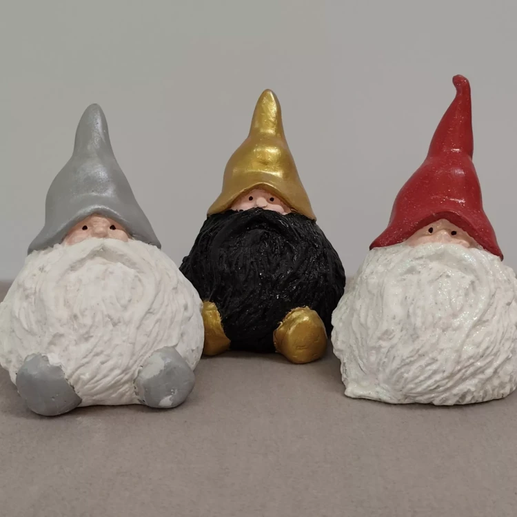Trzy figurki krasnali ustawione obok siebie na neutralnym tle: pierwszy ma srebrną czapkę i białą brodę, drugi złotą czapkę i czarną brodę, trzeci czerwoną czapkę i białą brodę. Wszystkie mają okrągłe nosy.