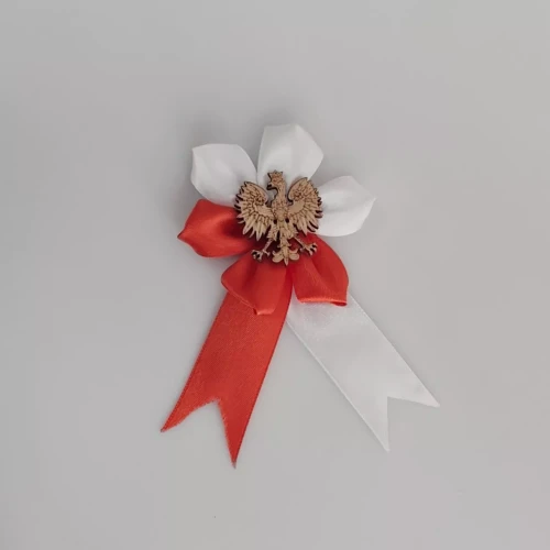 Kotylion patriotyczny z czerwono-białych wstążek ułożonych w kształt rozety, z drewnianym godłem Polski orłem w koronie pośrodku. Symbol narodowy używany podczas świąt i uroczystości upamiętniających polską historię i tożsamość.