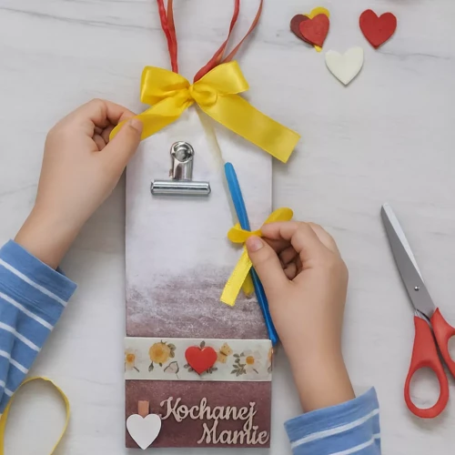 Dziecięce dłonie tworzą ręcznie robiony prezent z napisem „Kochanej Mamie”: ozdobna tabliczka z żółtą kokardą, serduszkami i przymocowanym długopisem. Na stole leżą nożyczki i dekoracje, scena pełna czułości i kreatywności.