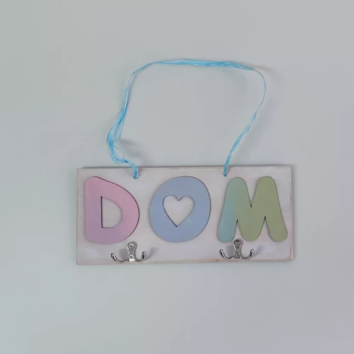 Dekoracyjna zawieszka ścienna z napisem „DOM” w kolorowych literach: różowe „D”, niebieskie „O” z sercem, zielone „M”. Litery na jasnej tabliczce z dwoma haczykami na dole i niebieską wstążką do zawieszenia. Połączenie ozdoby i funkcji.