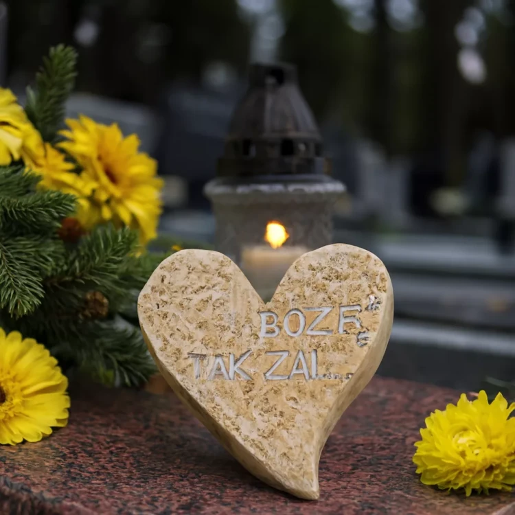 Gipsowe serce  z napisem „BOŻE TAK ŻAL…” leży na nagrobku wśród żółtych kwiatów i gałązek iglastych. W tle widoczny jest zapalony znicz w szklanym lampionie, scena pełna zadumy i pamięci o zmarłym.