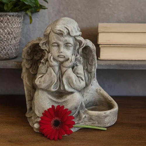 Gipsowa figurka siedzącego aniołka o zadumanym wyrazie twarzy, z podpartymi dłońmi i skrzyżowanymi nogami. Przed nim leży czerwona gerbera. W tle widać doniczkę z rośliną i stos książek na jasnej półce.