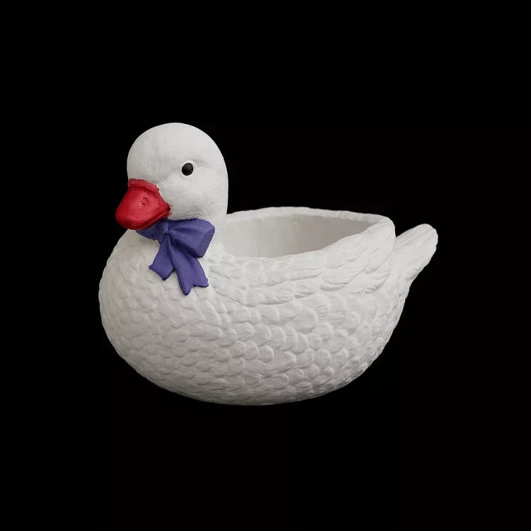 Biała gipsowa kaczka Kaja z fioletową kokardką na szyi, z fakturą piór i otwartym wnętrzem na drobne dekoracje, przedstawiona na czarnym tle.