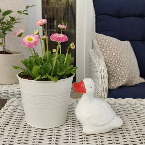 Białe wiklinowe meble z doniczką różowych kwiatów i białą figurką gęsi na stole; w tle fotel z granatową poduszką i rośliną w białej donicy, tworzące przytulny, dekoracyjny kącik.