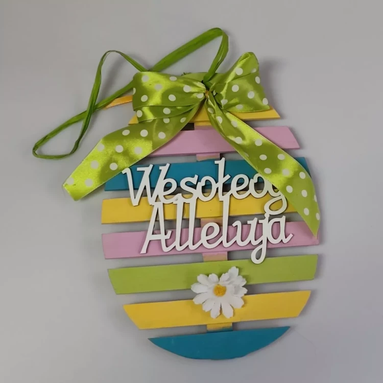 Drewniana pisanka z poziomych, pastelowych listewek, ozdobiona zieloną wstążką w groszki u góry, białym napisem „Wesołego Alleluja” na środku oraz białym kwiatkiem z żółtym środkiem na zielonej listewce.