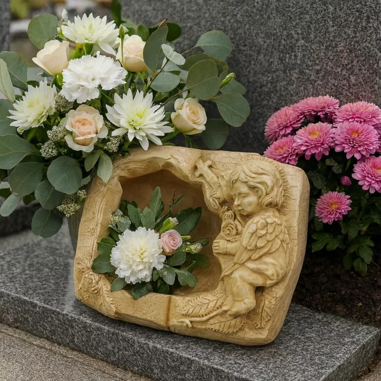 Gipsowa otwarta księgaz aniołkiem trzymającym krzyż i różę, ustawiona na nagrobku wśród białych, różowych i fioletowych kwiatów jako element dekoracji pamięci.