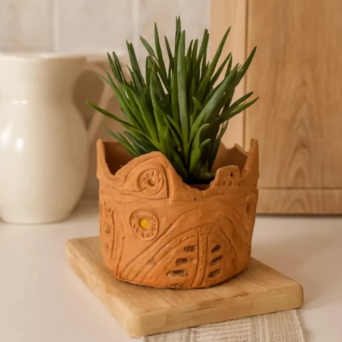 Osłonka ceramiczna w kolorze terakoty z rzeźbionymi detalami, ustawiona na drewnianej desce, z sukulentem w środku, w jasnym, domowym wnętrzu.