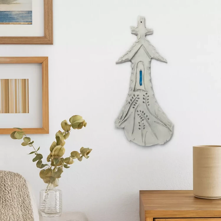 Ceramiczna kapliczka z niebieskim okienkiem zawieszona na białej ścianie w przytulnym wnętrzu. Obok znajdują się ramki ze sztuką, wazon z eukaliptusem, beżowy koc na fotelu i ceramiczny pojemnik na drewnianej komodzie.