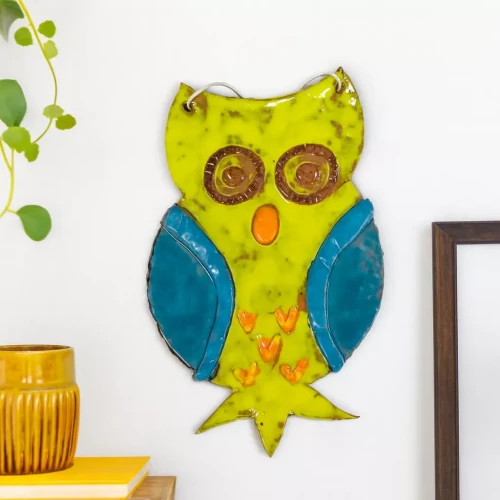 Ceramiczna sowa wisząca na białej ścianie, żółta z niebieskimi skrzydłami, pomarańczowym dziobem i serduszkami. W tle roślina, książki, żółte naczynie i drewniana ramka.