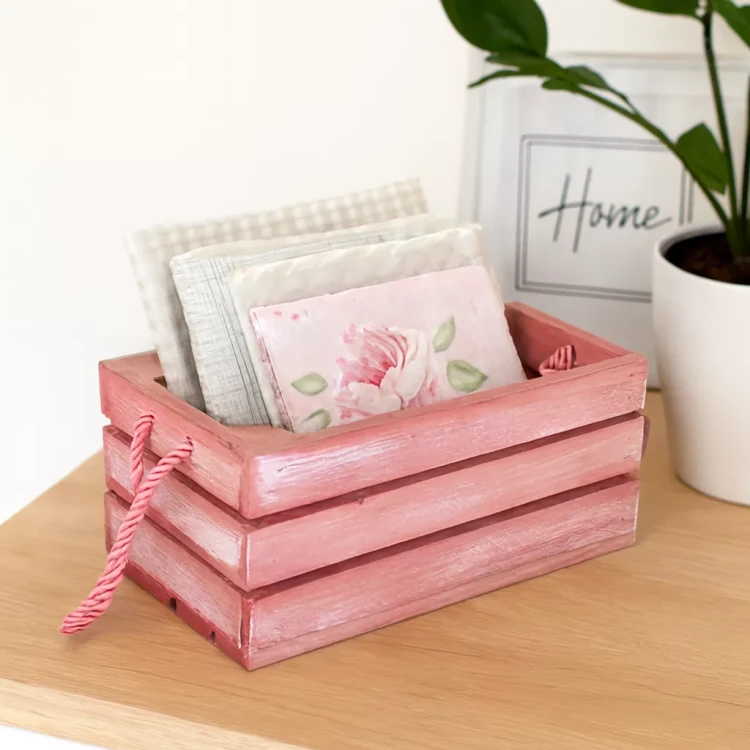Drewniana różowa skrzynka ze sznurkowymi uchwytami, wypełniona złożonymi serwetkami o kwiatowych i wzorzystych motywach, ustawiona na jasnym blacie obok doniczki z rośliną i dekoracyjnej ramki.