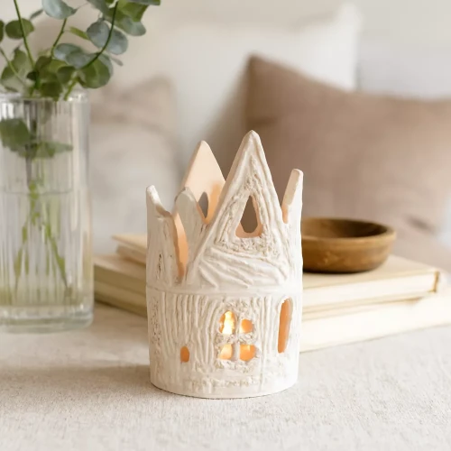 Ceramiczny lampion w kształcie bajkowego domku z fantazyjnymi dachami i okienkami, ustawiony w domowym wnętrzu na tle jasnych poduszek i dekoracji, oświetlony naturalnym światłem dziennym.