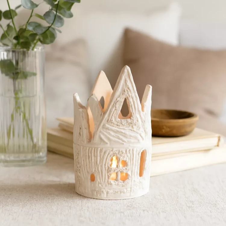 Ceramiczny lampion w kształcie bajkowego domku z fantazyjnymi dachami i okienkami, ustawiony w domowym wnętrzu na tle jasnych poduszek i dekoracji, oświetlony naturalnym światłem dziennym.