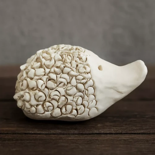 Ceramiczna figurka jeża o gładkim pyszczku i dwóch małych otworach jako oczy, z grzbietem pokrytym licznymi spiralnymi wypustkami przypominającymi kolce, ustawiona na drewnianej powierzchni.