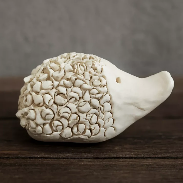 Ceramiczna figurka jeża o gładkim pyszczku i dwóch małych otworach jako oczy, z grzbietem pokrytym licznymi spiralnymi wypustkami przypominającymi kolce, ustawiona na drewnianej powierzchni.