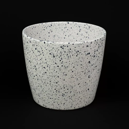 Biała osłonka ceramiczna o cylindrycznym kształcie, pokryta drobnymi, nieregularnymi granatowo  niebieskimi kropkami, stojąca na czarnym tle.