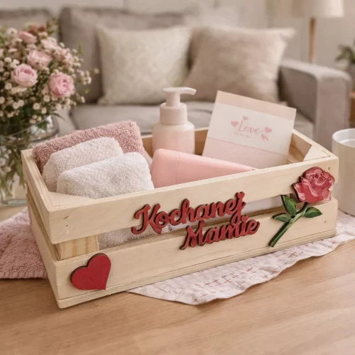Drewniana skrzynka prezentowa z napisem „Kochanej Mamie”, wypełniona ręcznikami, kosmetykami i kartką „Love”, ustawiona w przytulnym wnętrzu z kwiatami w tle.