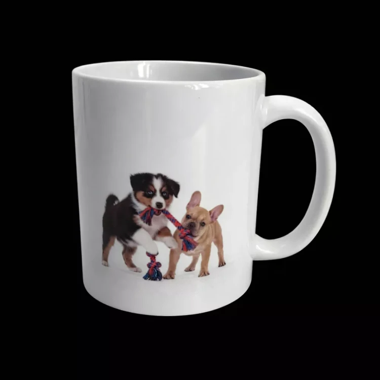 Biały ceramiczny kubek z nadrukiem dwóch szczeniaków bawiących się szarpakiem, czarno-brązowy i jasny pies ciągną linową zabawkę. Kubek na czarnym tle.