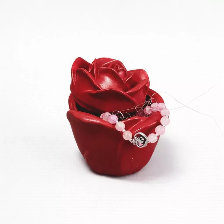 Czerwona dekoracyjna róża gipsowa, na której ułożona jest różowa bransoletka z koralików, leżąca na białym tle.