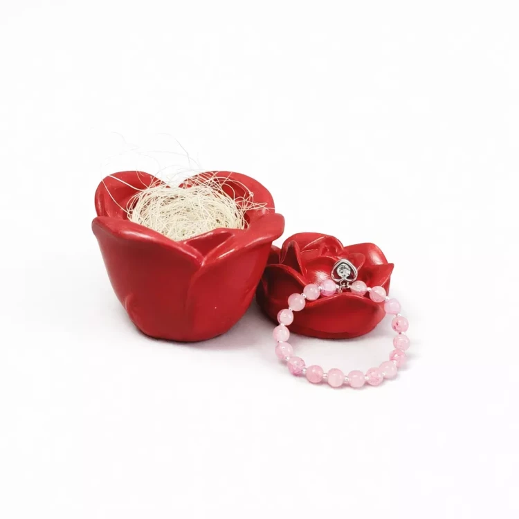 Gipsowe pudełeczko w kształcie czerwonej róży; jedno otwarte z jasnym wypełnieniem, pokrywka pudełeczka z różową bransoletką z serduszkiem ułożoną na wierzchu, na białym tle.
