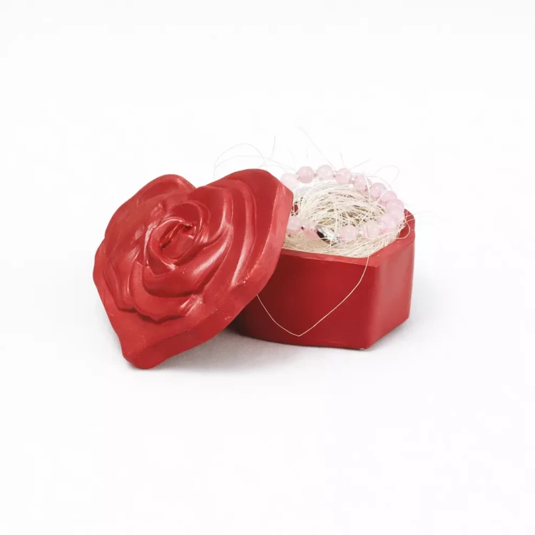 Gipsowe pudełeczko pomalowane na czerwono w kształcie serca z różaną pokrywką, częściowo otwarte, w środku różowa bransoletka na jasnym wypełnieniu, na białym tle.