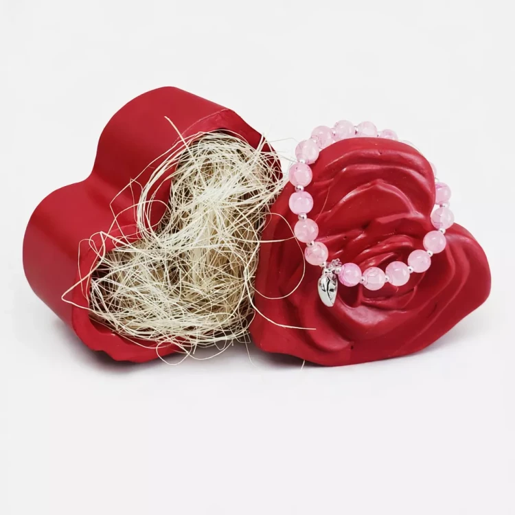 Czerwone gipsowe pudełeczko w kształcie serca z fakturą różanych płatków, otwarte i wypełnione jasnym sizalem, z różową bransoletką z koralików i srebrnym listkiem ułożoną na brzegu.
