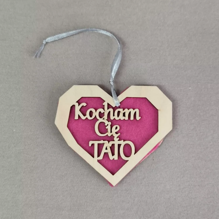 Drewniana ozdoba w kształcie serca z różowym tłem filcowym i napisem ‘Kocham Cię Tato’, zawieszona na srebrnej wstążce.