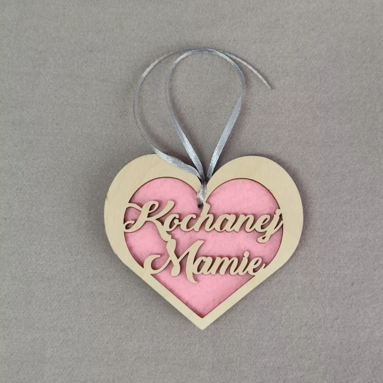 Zawieszka w kształcie serca z drewnianą ramką i różowym tłem z filcu, z wyciętym napisem ‘Kochanej Mamie’, zawieszona na srebrnej wstążce.