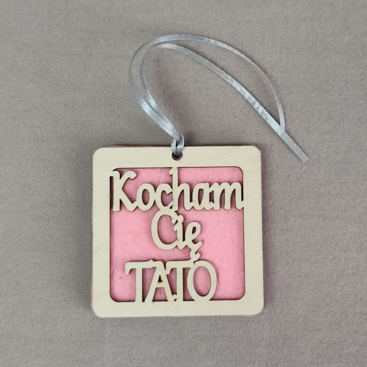 Kwadratowa drewniana zawieszka z różowym filcowym tłem i wyciętym napisem ‘Kocham Cię Tato’, ozdobiona srebrną wstążką do powieszenia.