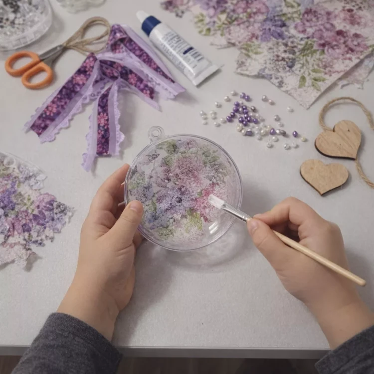 Dziecięce dłonie ozdabiają akrylowy medalion techniką decoupage; na stole leżą nożyczki, klej, wstążki, koraliki i serduszka z napisem „Kochanej Mamie”.