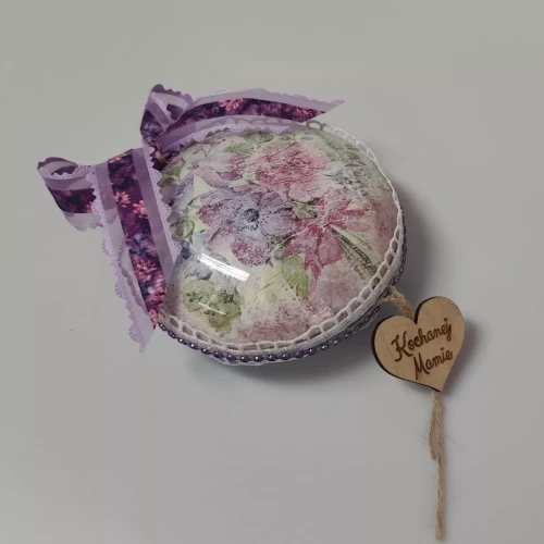 Ręcznie zdobiona, kopułowa ozdoba w pastelowych kolorach z kwiatowym motywem, koronką i perełkami, przewiązana fioletową wstążką; na drewnianym serduszku widnieje napis "Kochanej Mamie"