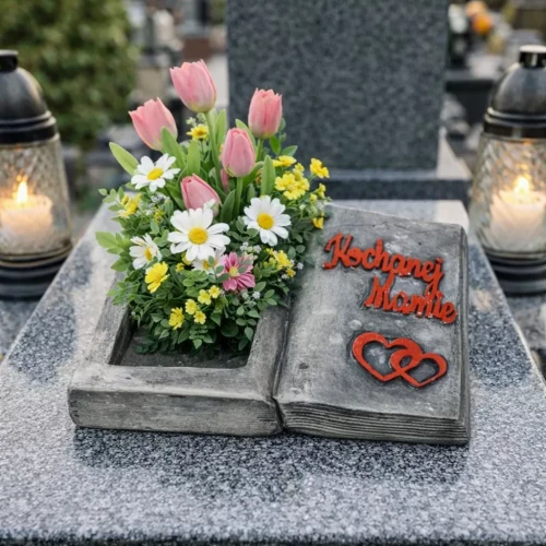 Osłonka gipsowa w kształcie otwartej książki z napisem „Kochanej Mamie” i czerwonymi sercami, leżąca na nagrobku. W zagłębieniu po lewej stronie sztuczne wiosenne kwiaty — tulipany, stokrotki i żółte drobne kwiatki.