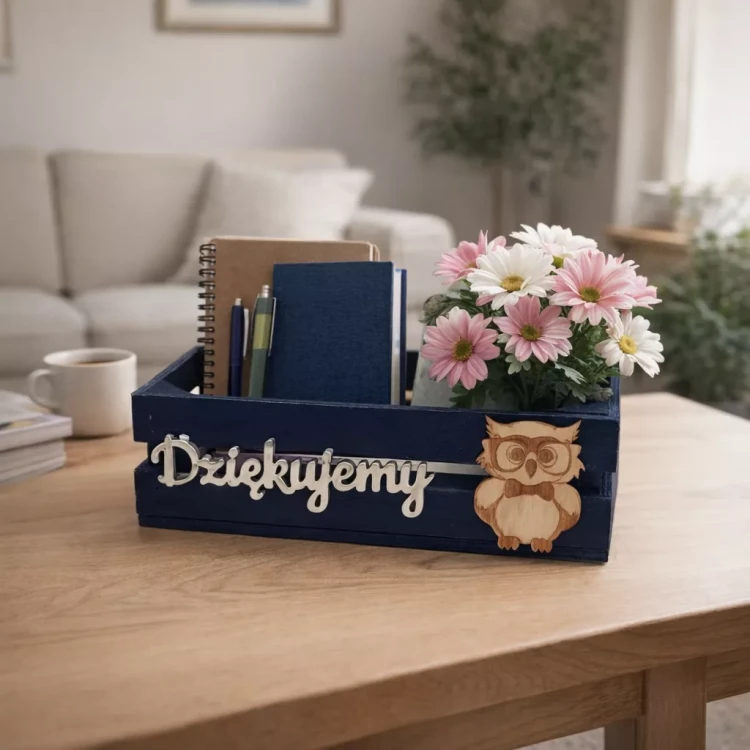 Granatowa drewniana skrzynka z napisem „Dziękujemy” i małą sówką, wypełniona zeszytami, długopisami i bukietem różowo‑białych stokrotek, ustawiona na stole w przytulnym salonie.