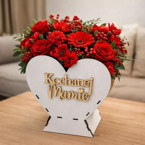 Aranżacja czerwonych kwiatów róż, gerber ułożonych w białej, sercowatej osłonce z drewnianym napisem „Kochanej Mamie”, stojąca na drewnianym stole w domowym wnętrzu.
