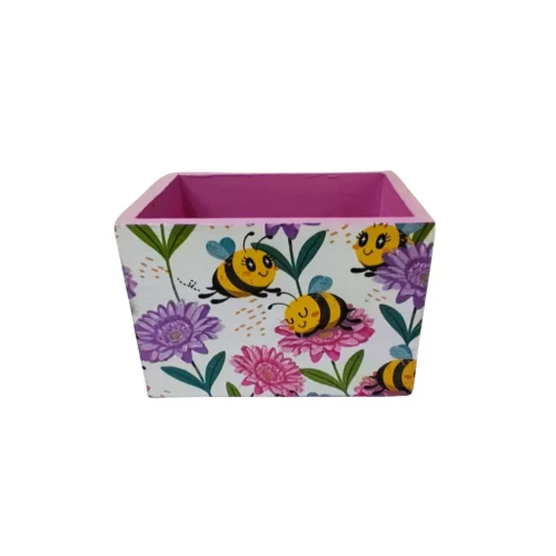 Drewniane pudełko z jasnoróżowym wnętrzem i białymi bokami ozdobionymi rysunkami uśmiechniętych pszczół oraz barwnych kwiatów w różu, fiolecie i zieleni. Lekka, pogodna dekoracja do przechowywania drobiazgów.