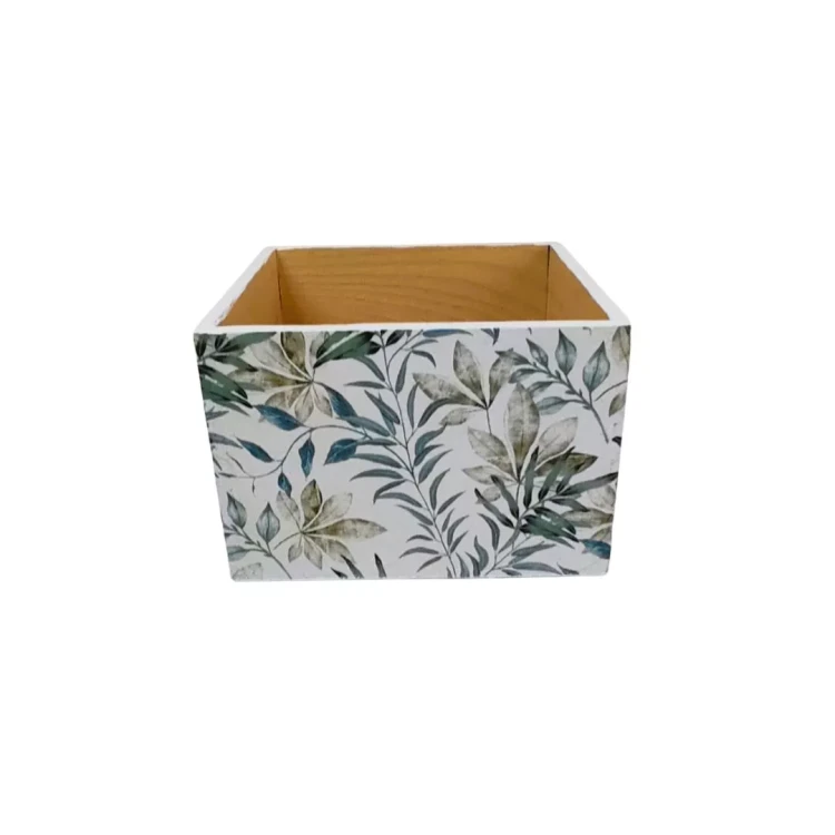 Otwarta skrzynka z jasnego drewna, z zewnętrzną stroną pokrytą dekoracyjnymi liśćmi. Zieleń na białym tle nadaje jej lekkość. Wnętrze surowe i minimalistyczne.