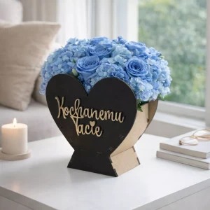 Drewniany pojemnik w kształcie serca z napisem „Kochanemu Tacie” na froncie z czarnej pleksi, wypełniony niebieskimi kwiatami, stoi na stole w jasnym salonie przy oknie. W tle widać kanapę, książki i świecę w świetle dziennym.