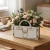 Biały flower box w kształcie torebki z ozdobnym grawerem, stojący na drewnianym stole. Wypełniony jest obfitym bukietem różowych róż, fioletowej lawendy, rumianków i eukaliptusa. W tle widać kubek, serwetkę i książki w jasnym pomieszczeniu.