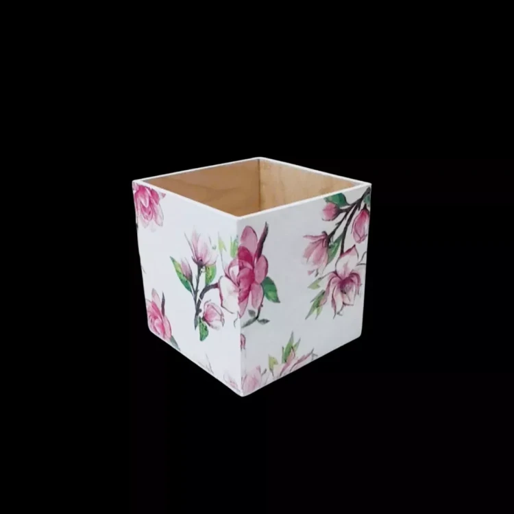 Białe kwadratowe pudełko z drewna ozdobione techniką decoupage. Na ściankach widnieje wzór gałązek z różowymi kwiatami magnolii. Wnętrze pudełka jest w kolorze naturalnego, jasnego drewna. Tło czarne, produkt pokazany pod kątem.