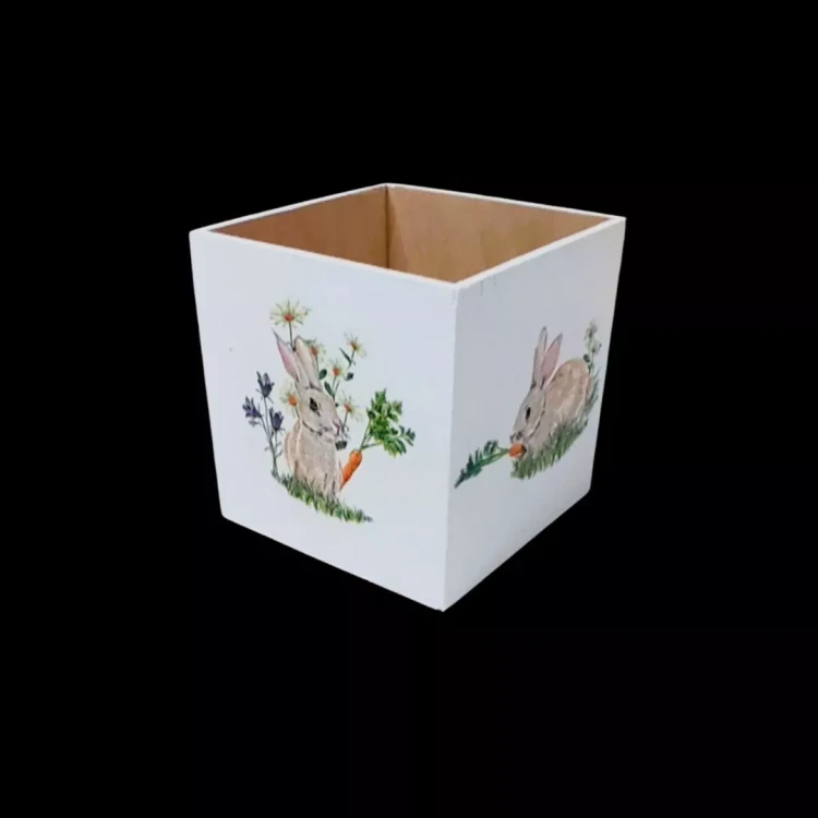 Drewniany pojemnik pomalowany na biało z akwarelowymi grafikami szarych zajączków. Jeden królik siedzi przy niebieskich kwiatach, drugi je marchewkę. Estetyka rustykalna i wiosenna.