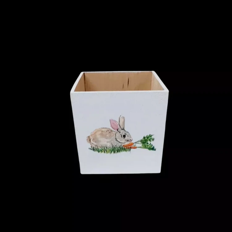 Kwadratowe pudełko drewniane, białe, technika decoupage. Motyw: króliki, kwiaty, marchewki. Widok z przodu ukazuje ściankę i naturalne, drewniane wnętrze.