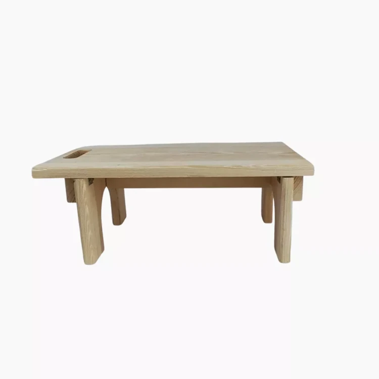 Składany drewniany stołeczek lub podnóżek z jasnego naturalnego drewna. Prostokątny blat z zaokrąglonymi krawędziami i wyciętym uchwytem po lewej stronie. Cztery składane nogi. Produkt sfotografowany na jasnym, jednolitym tle.